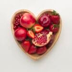 Heart-shaped bowl filled with aphrodisiac fruits like strawberries, pomegranate, and red apples on white background